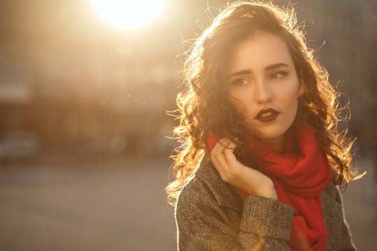 Street portrait of lovely brunette girl with red lips walking down the city with sun glare. Empty space