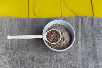 bowl of flax flour, a spoon with flax seeds on a yellow wooden background and linen canvas