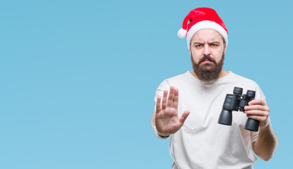 Young caucasian hipster man wearing christmas hat looking though binoculars over isolated background with open hand doing stop sign with serious and confident expression, defense gesture