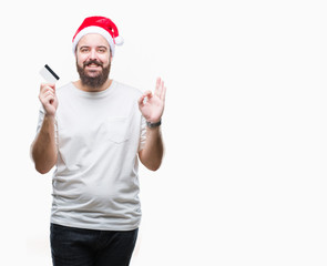 Young caucasian man wearing christmas hat holding credit card over isolated background doing ok sign with fingers, excellent symbol