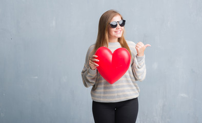 Young adult woman over grey grunge wall holding red heart in love pointing and showing with thumb up to the side with happy face smiling