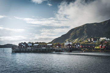 Rainbow ofer red houses rorbuer of Reine in Lofoten, Norway with red rorbu houses, clouds, rainy blue sky and sunny. Bridges and mountains in the background.