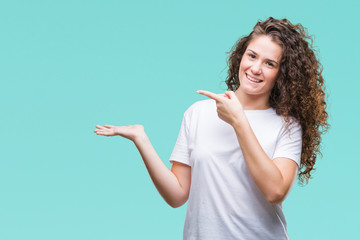 Beautiful brunette curly hair young girl wearing casual t-shirt over isolated background amazed and smiling to the camera while presenting with hand and pointing with finger.