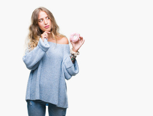 Beautiful young blonde woman eating pink donut over isolated background serious face thinking about...