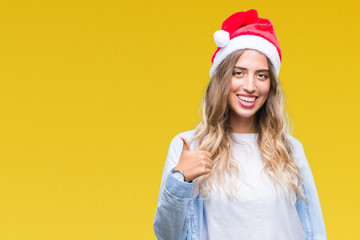 Beautiful young blonde woman wearing christmas hat over isolated background doing happy thumbs up gesture with hand. Approving expression looking at the camera with showing success.
