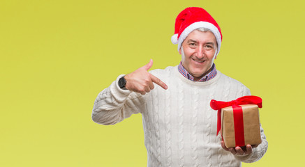 Handsome senior man wearing christmas hat and holding gift over isolated background with surprise face pointing finger to himself
