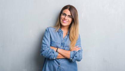 Young adult woman over grunge grey wall wearing glasses happy face smiling with crossed arms...