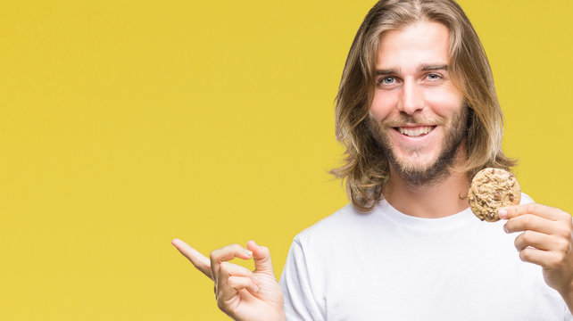 Young handsome man with long hair eating chocolate cooky over isolated background very happy pointing with hand and finger to the side