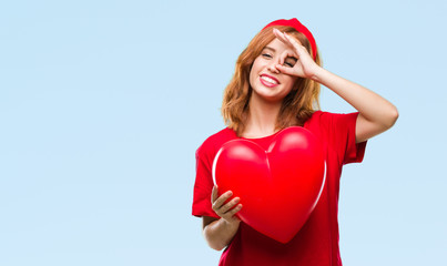 Young beautiful woman holding red heart in love over isolated background with happy face smiling doing ok sign with hand on eye looking through fingers
