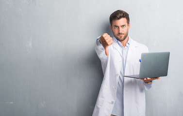 Handsome young man over grey grunge wall wearing white coat using laptop with angry face, negative...