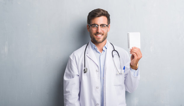 Handsome young doctor man over grey grunge wall holding blank notebook with a happy face standing and smiling with a confident smile showing teeth