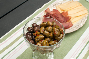 Glass bowl of olives in the olive oil and saucer with pastrami and cheese as appetizers closeup on covered with textile mat home table