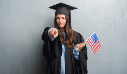 Young brunette woman over grunge grey wall wearing graduate uniform holding flag of America pointing with finger to the camera and to you, hand sign, positive and confident gesture from the front