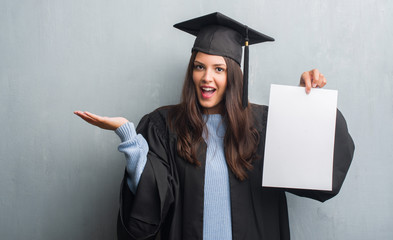 Young brunette woman over grunge grey wall wearing graduate uniform holding degree very happy and...