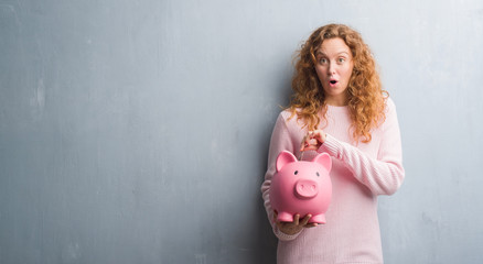 Young redhead woman over grey grunge wall holding piggy bank and dollar scared in shock with a...