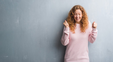 Young redhead woman over grey grunge wall wearing pink sweater excited for success with arms raised celebrating victory smiling. Winner concept.