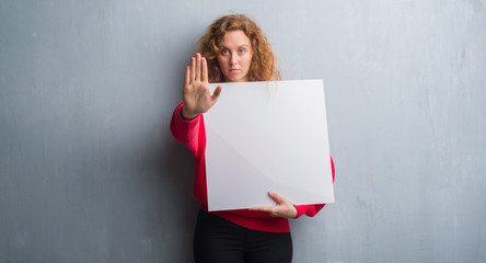 Young redhead woman over grey grunge wall holding advertising banner with open hand doing stop sign with serious and confident expression, defense gesture