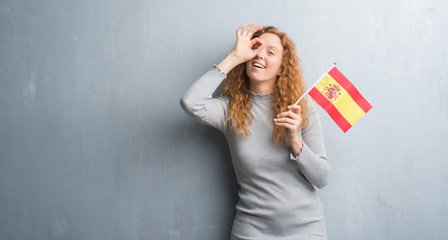 Young redhead woman over grey grunge wall holding flag of Spain with happy face smiling doing ok sign with hand on eye looking through fingers