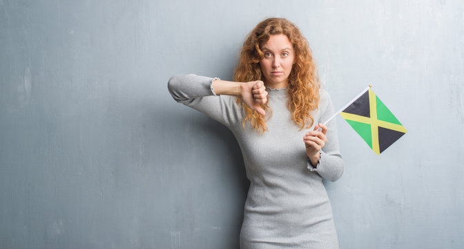 Young redhead woman over grey grunge wall holding flag of Jamaica with angry face, negative sign showing dislike with thumbs down, rejection concept