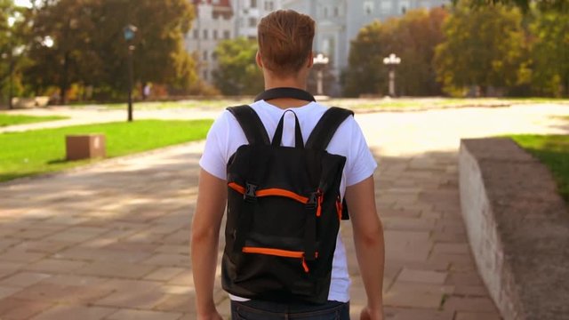 back view young man walking with backpack on the street. caucasian student with blond hair walks outdoor slow motion