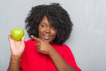 Young african american woman over grey grunge wall eating green apple very happy pointing with hand and finger