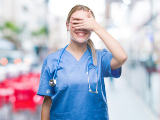 Young blonde surgeon doctor woman over isolated background smiling and laughing with hand on face covering eyes for surprise. Blind concept.