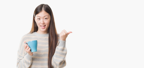 Young Chinese woman over isolated background driking cup of coffee pointing and showing with thumb up to the side with happy face smiling