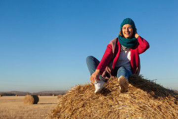 Naklejka na ściany i meble A young woman in a blue knitted hat, pink coat and jeans enjoys nature, sitting on a haystack around blue sky. The concept of livestyle and outdoor recreation in autumn