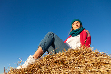 Naklejka na ściany i meble Fashion lifestyle portrait of young trendy woman dressed in pink coat, pink knitted hat posing, laughing and smiling on a haystack around blue sky. portrait of joyful woman
