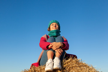 Young attractive lady in blue knitting hat and pink coat sitting on haystack. Smiling beautiful...