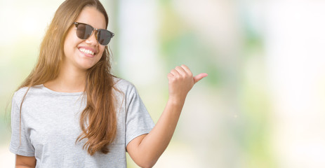 Young beautiful brunette woman wearing sunglasses over isolated background smiling with happy face looking and pointing to the side with thumb up.