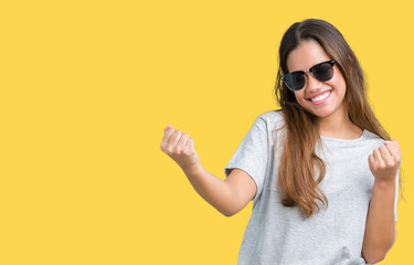 Fototapeta premium Young beautiful brunette woman wearing sunglasses over isolated background celebrating surprised and amazed for success with arms raised and open eyes. Winner concept.