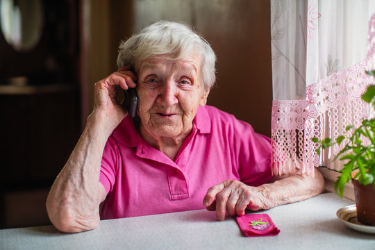Elderly woman talking on a mobile phone sitting at a table in his house.