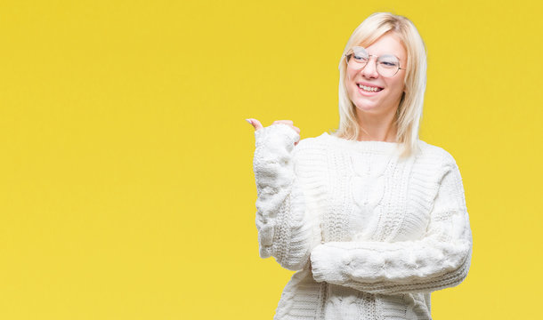 Young beautiful blonde woman wearing winter sweater and glasses over isolated background smiling with happy face looking and pointing to the side with thumb up.