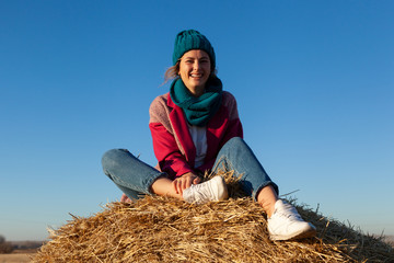 Naklejka na ściany i meble Portrait female student on field background. Beautiful modern young woman wearing pink knitting hat smile, look at the camera and enjoy autumn sunny day and sitting on haystick.