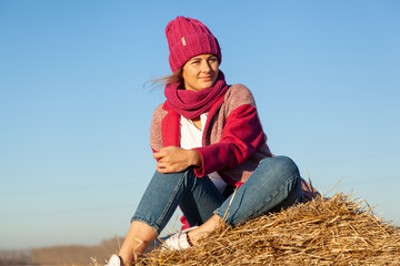 Naklejka na ściany i meble The concept of livestyle outdoor in autumn. Close up of a young woman student in a warm autumn clothes looking funny, smilling, posing for the camera on haystick