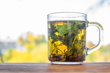 A cup of mint tea before autumn window with yellow trees on the background. Closeup, selective focus