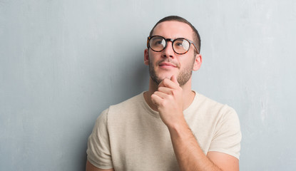 Young caucasian man over grey grunge wall serious face thinking about question, very confused idea