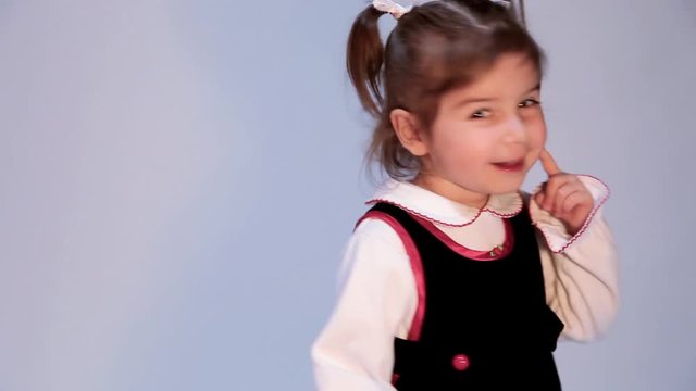 A cute, mischievous girl on a plain background bites her finger like she is thinking and turns to look behind her as if she expects a surprise.  There is copy space.