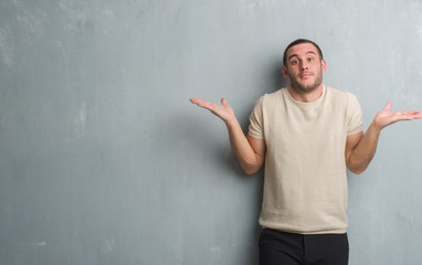 Young caucasian man over grey grunge wall clueless and confused expression with arms and hands...