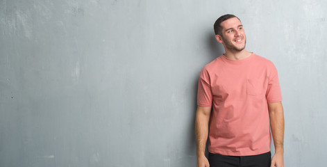 Young caucasian man over grey grunge wall looking away to side with smile on face, natural expression. Laughing confident.