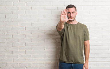 Young caucasian man standing over white brick wall doing stop sing with palm of the hand. Warning...