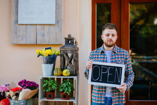 Young business man owner floral shop with open sign waiting for buyers near storefront outdoor in sunny autumn day.