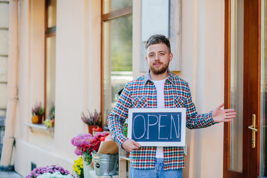 Cheerful, nice man owner floral store in informal wear holding opem sign, having his hands open like hospitable gesture, you are welcome, well met, standing over storefront of flower shop, outdoor.