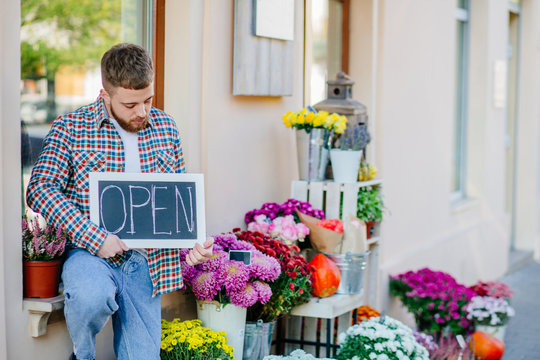 Young florist man holding sign open outdoor of flower shop near bouquets, autumn decoration, pumpkins. Concept of hospitable private business floristic art.