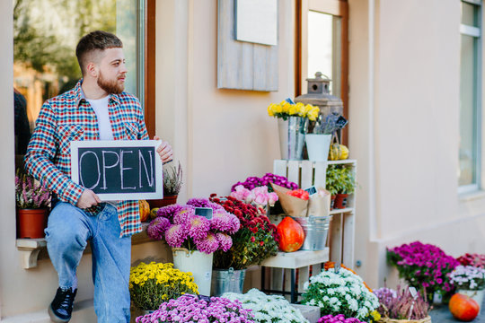 Young florist man holding sign open outdoor of flower shop near bouquets, autumn decoration, pumpkin.s. Concept of hospitable private business floristic art.