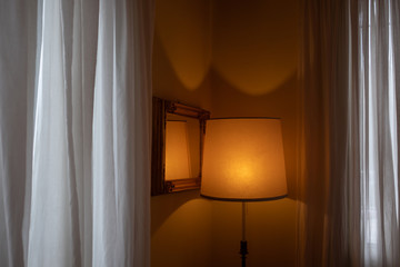 Empty elegant vintage hotel room at twilight with copy space. Orange lit abat-jour, framed mirror and white curtains