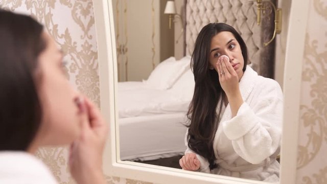 Young brown-haired women reflection in the mirror. Sitting back to the camera. Finishes her face make-up foundation and touches her forehead, right cheek, chin, cheek with a cotton sponge