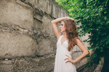 Beautiful moody girl with curly natural hair in white dress near green tree leaves. Summer beauty portrait. Sad lady in nature outdoors. Alone serious woman with pensive sight in long tunnel in forest