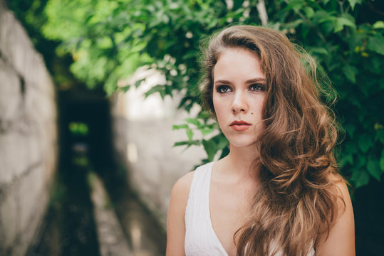 Beautiful moody girl with curly natural hair in white dress near green tree leaves. Summer beauty portrait. Sad lady in nature outdoors. Alone serious woman with pensive sight in long tunnel in forest